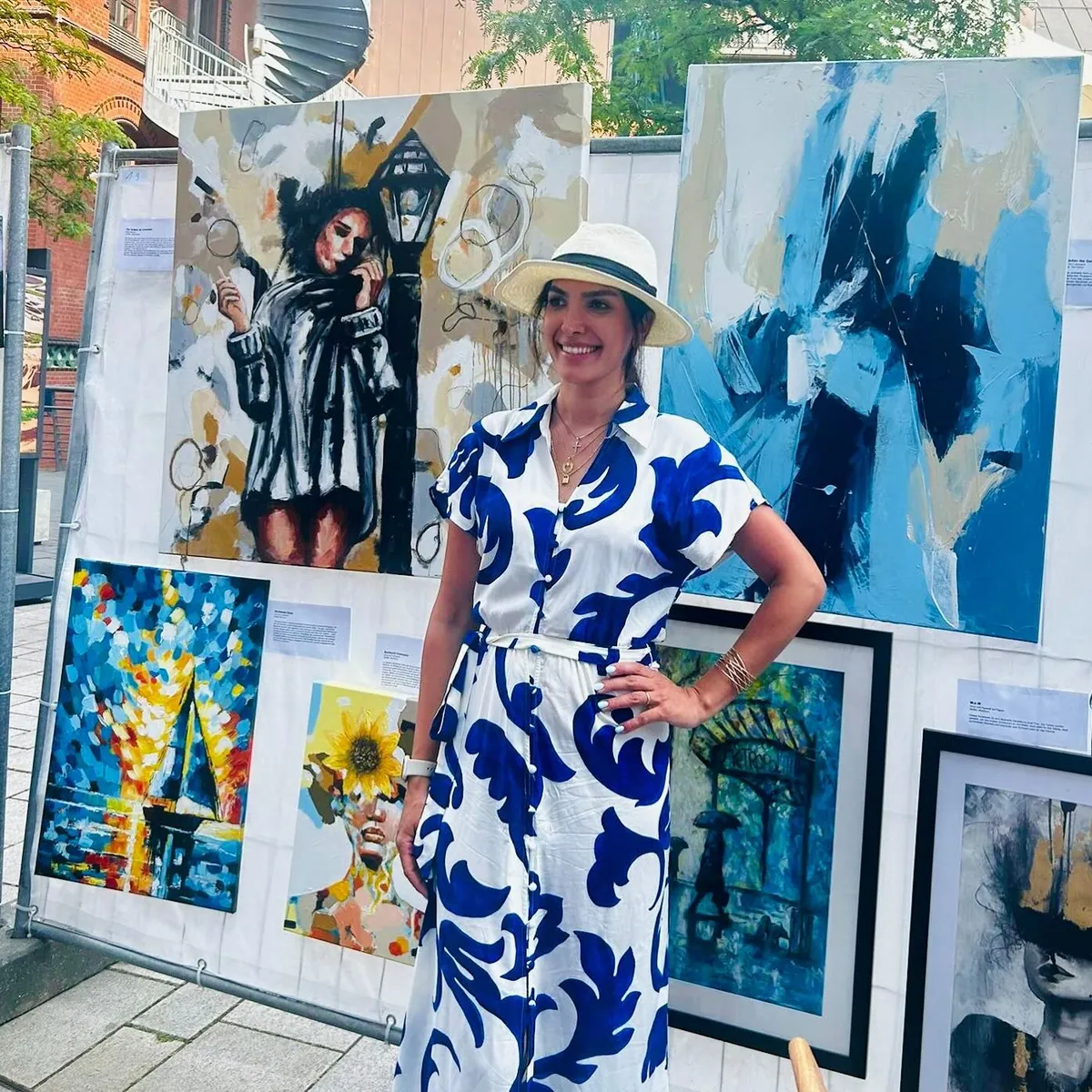 Pegah Koch in a white and blue dress and a straw hat, standing in front of her paintings at an open-air exhibition.